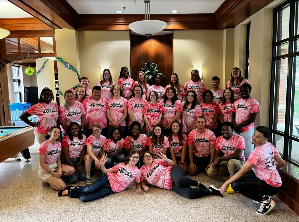 Group photo of res life staff wearing matching pink and white tie-dye t-shirts, gathered in a brightly lit indoor space.